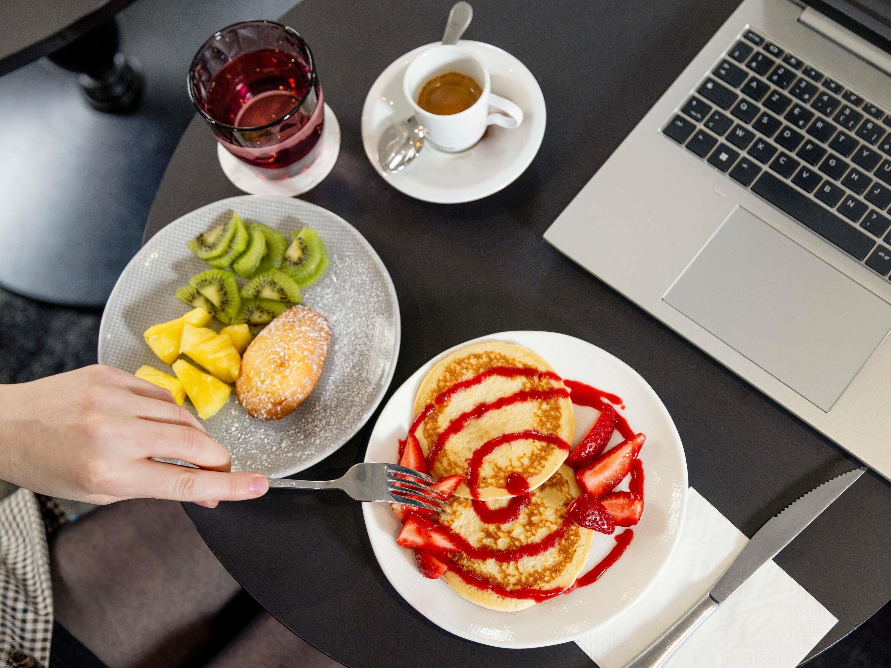 A meal with pancakes, kiwi, pineapple, and a beverage on a table with a laptop.