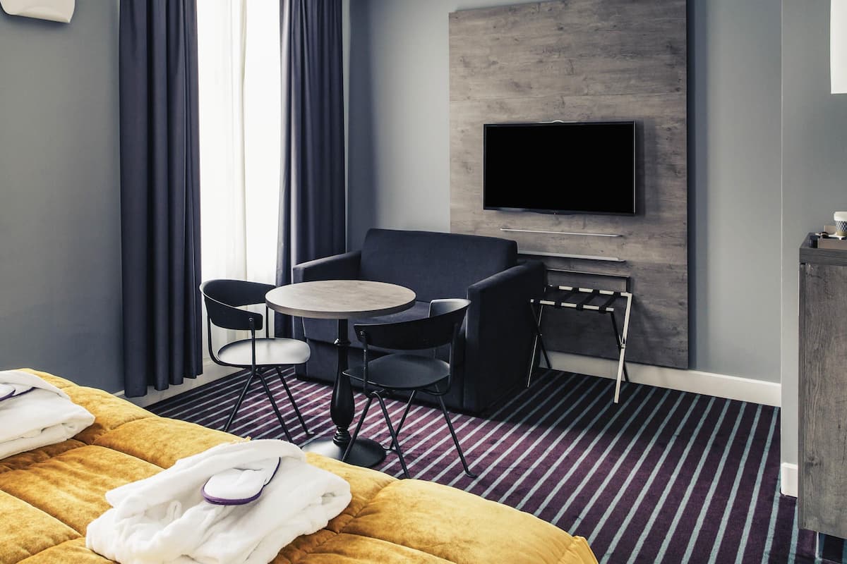 A hotel room with a bed, a round table with two chairs, a flat-screen TV mounted on a wooden panel, and a striped carpet.