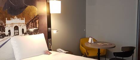 A hotel room with a bed, a desk, a chair, and a wall-mounted lamp.