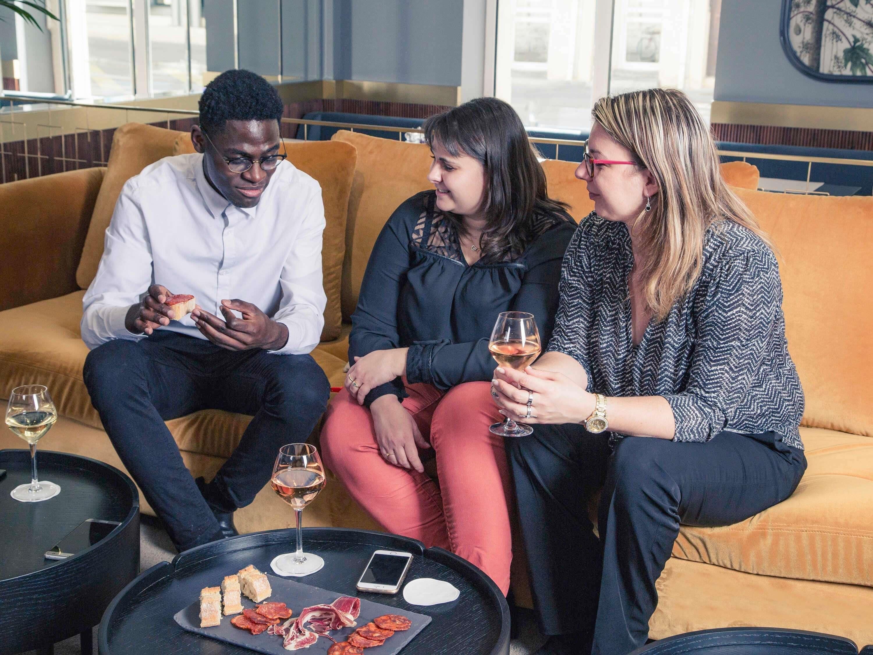 Three people seated on a sofa, a glass of wine, and a plate of food on a table.