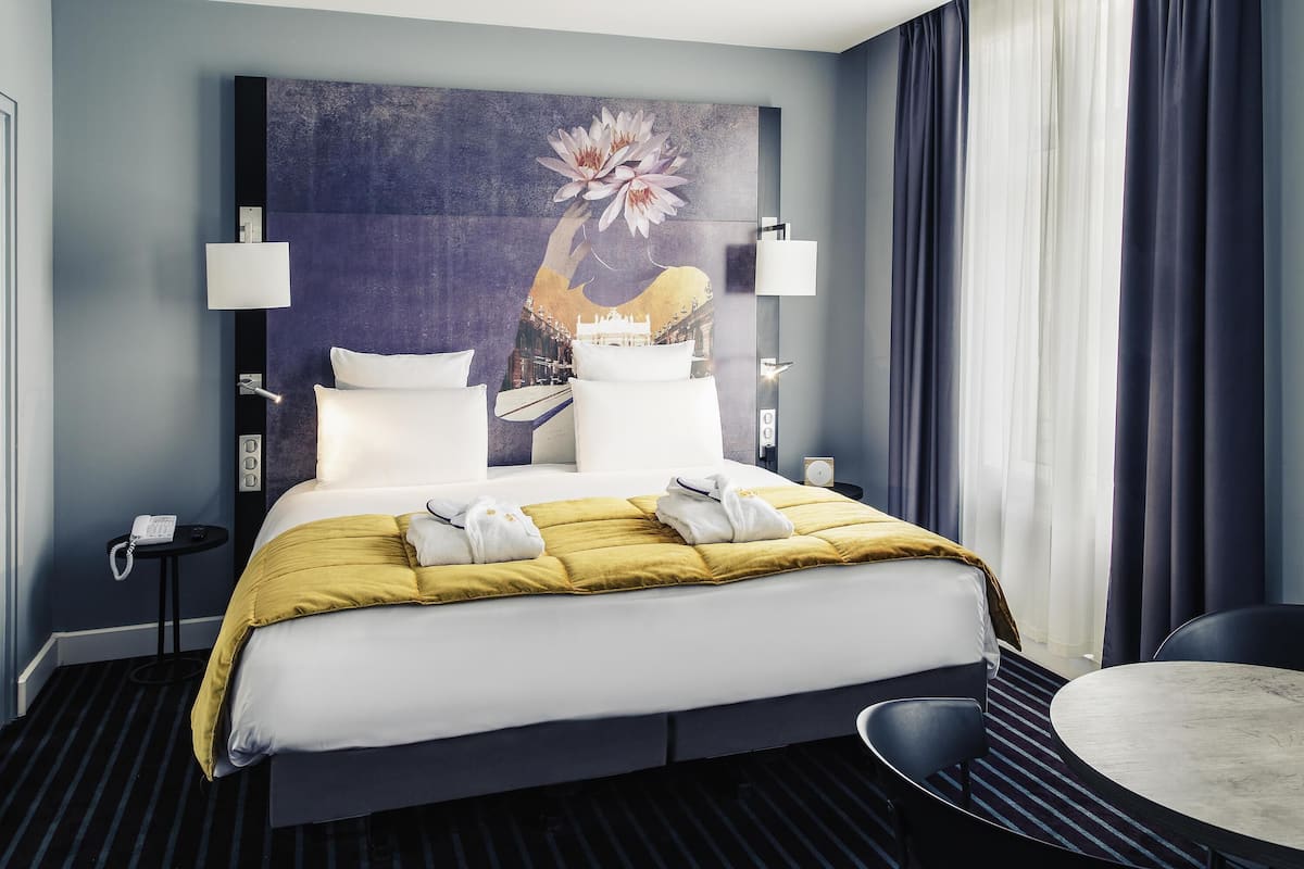 A hotel room with a bed, bedside lamps, a painting, a telephone, a chair, and a round table.