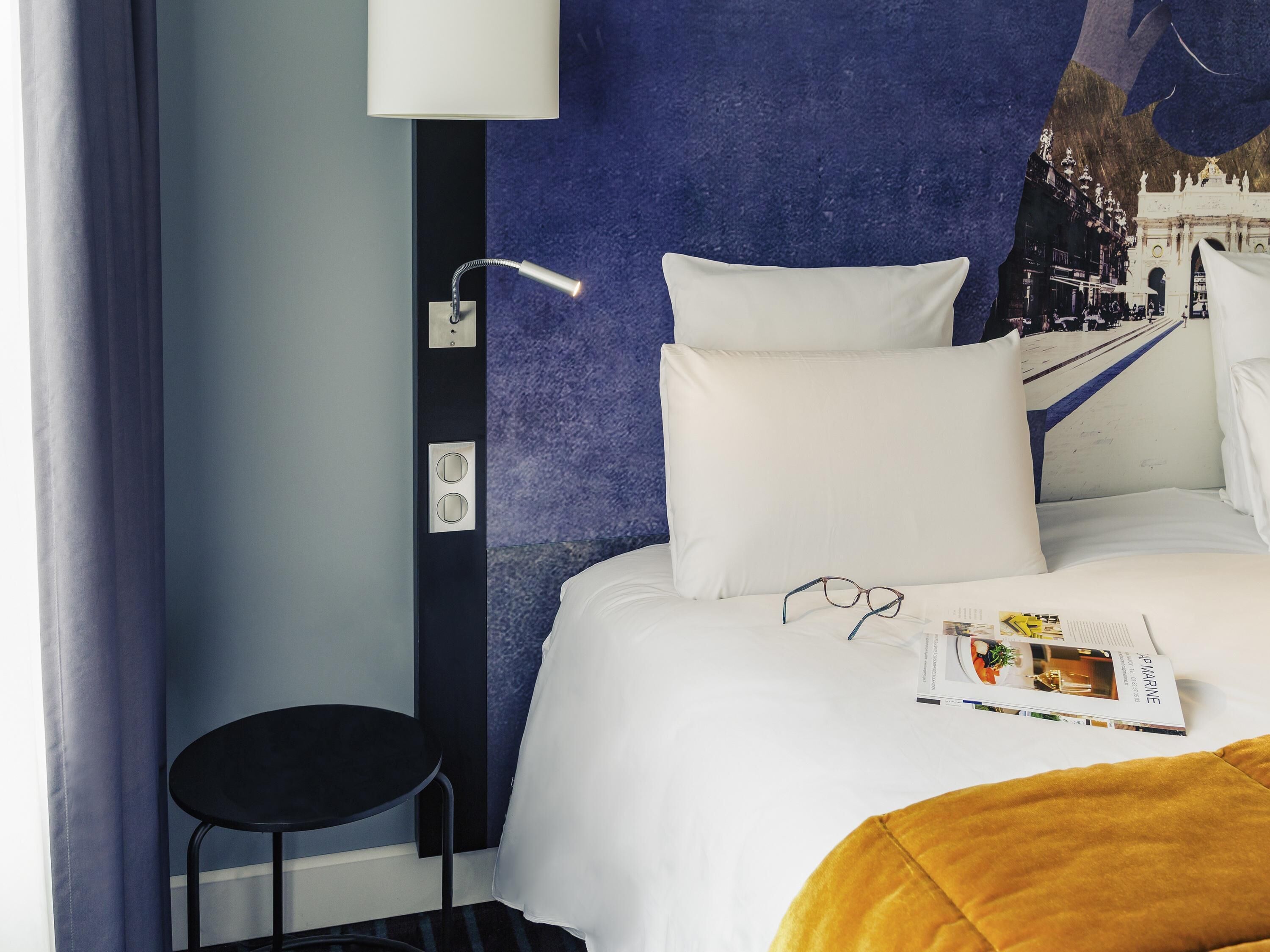 A hotel room with a bed, a black chair, a white pillow, and a magazine on the bed.