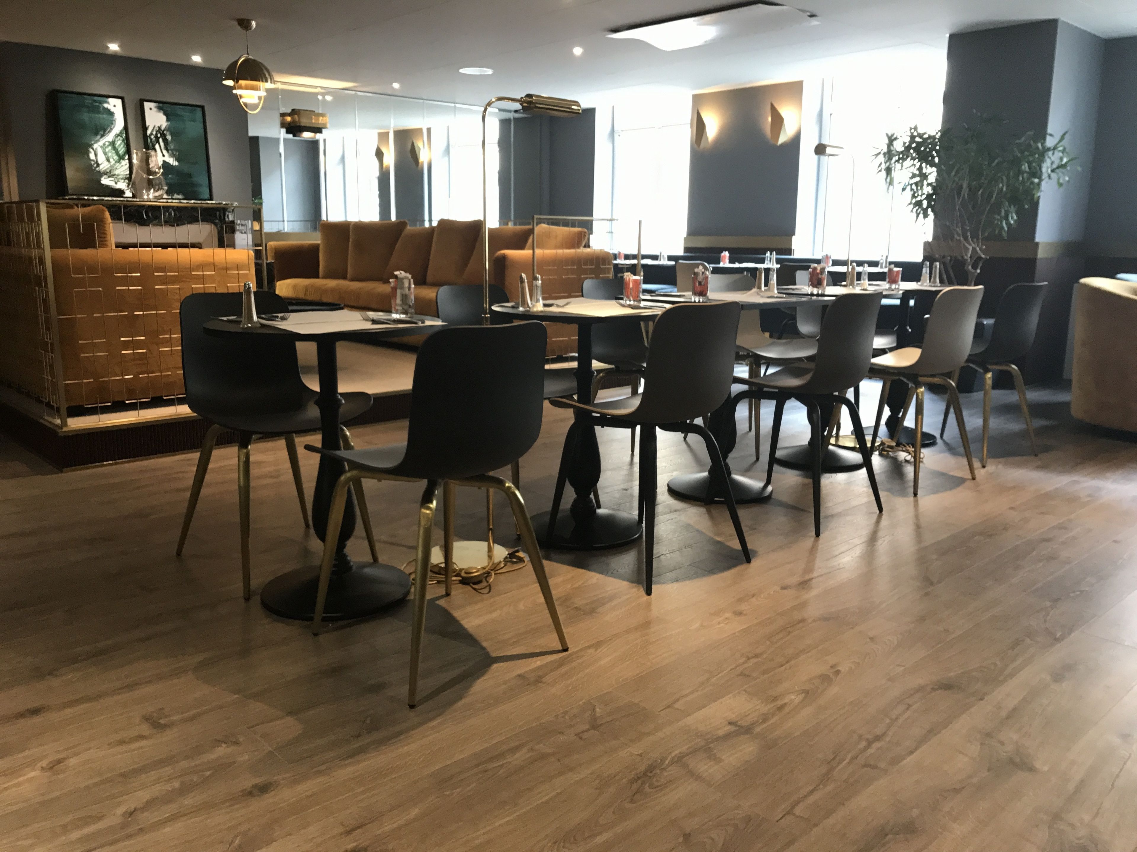 A modern dining area with wooden flooring, black and grey chairs, and tables. There are abstract paintings on the walls and a large window providing natural light.