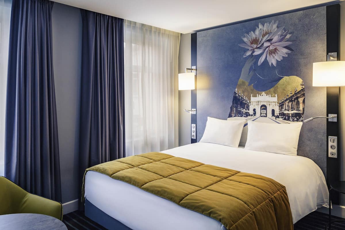 A hotel room with a large bed, a chair, a table, and a wall with a floral painting.