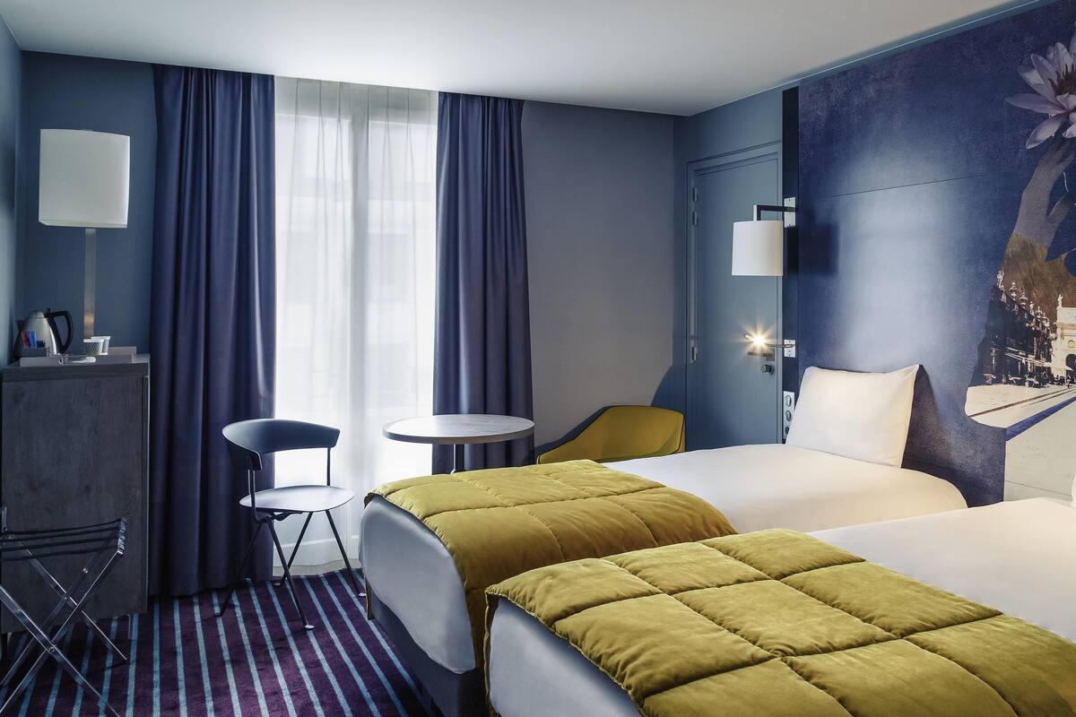 A hotel room with two beds, a desk, a chair, a small table, and a window with curtains.