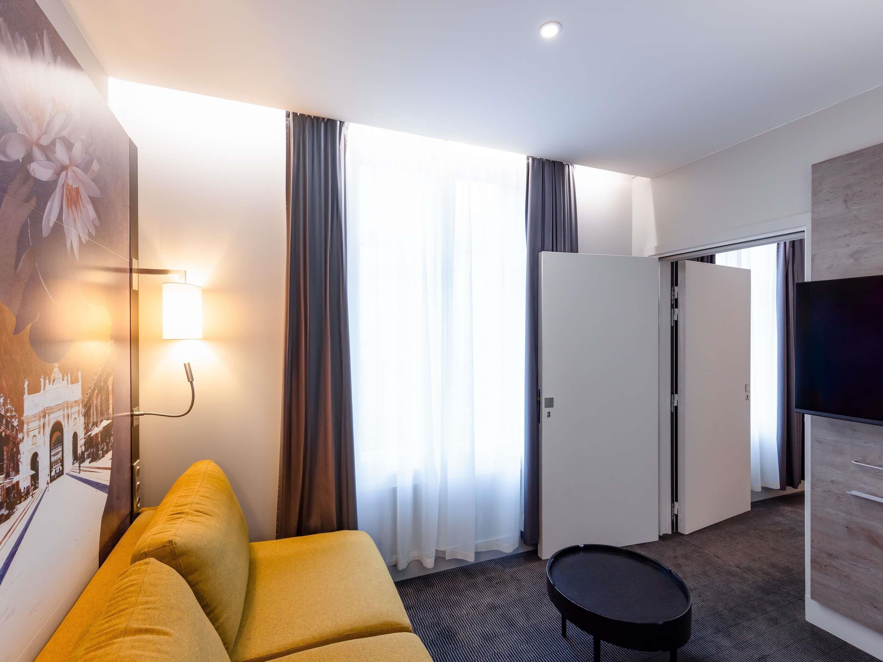 Habitación de hotel con sofá, una mesita, televisor, puerta y ventana con cortinas.