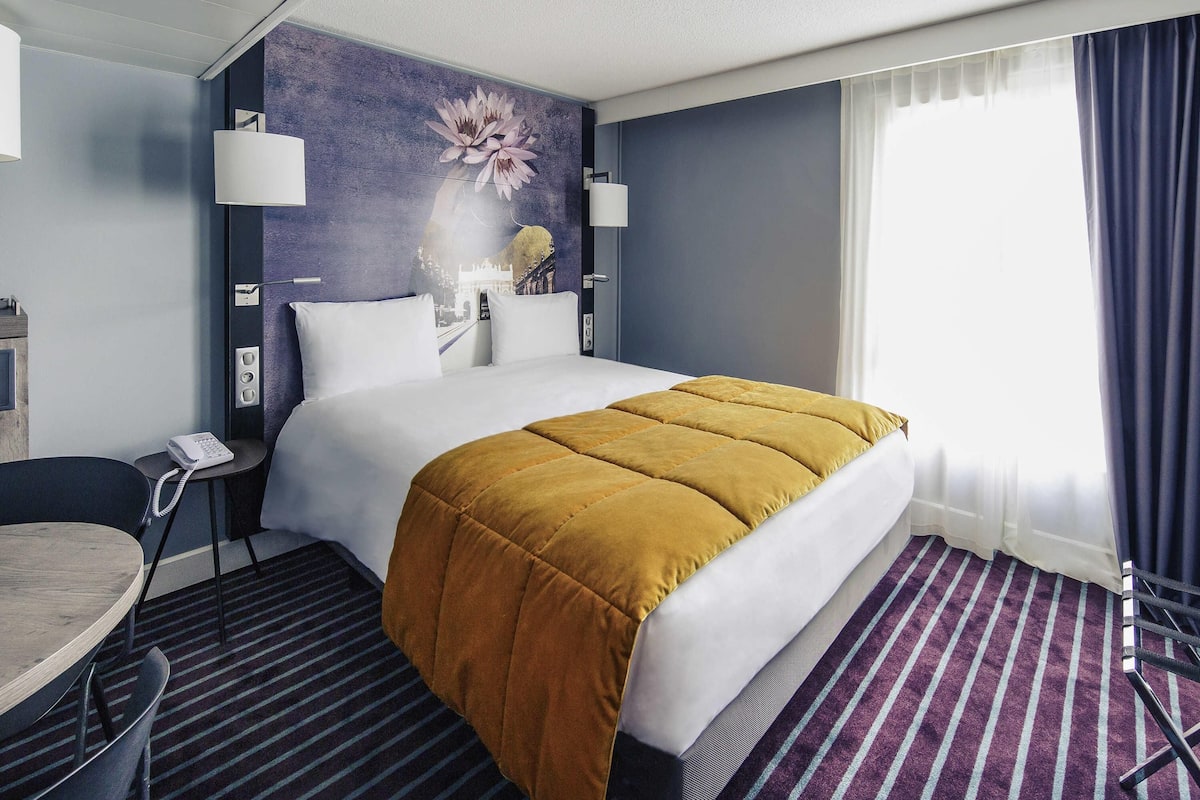 A hotel room with a bed, a desk, a telephone, and a wall with a floral mural.
