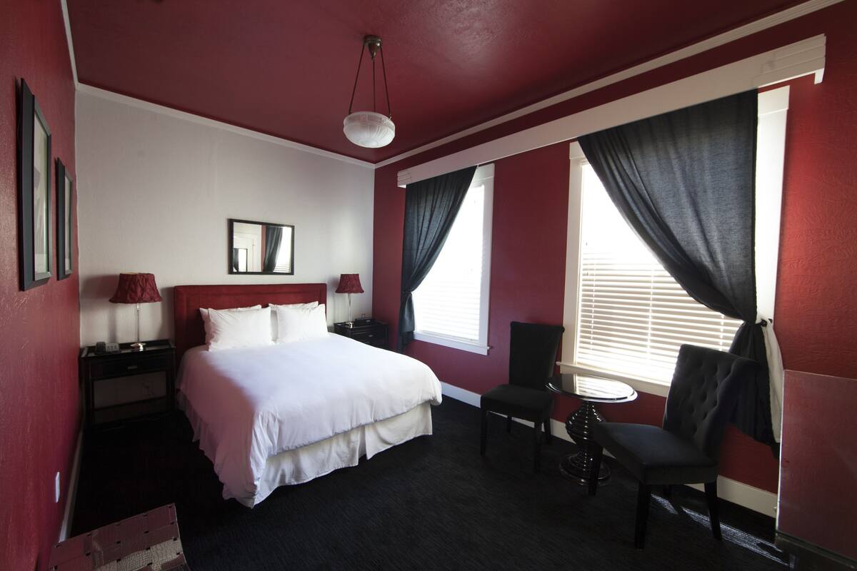 A hotel room with a bed, a chair, a small table, a nightstand, a lamp, and a window with curtains.