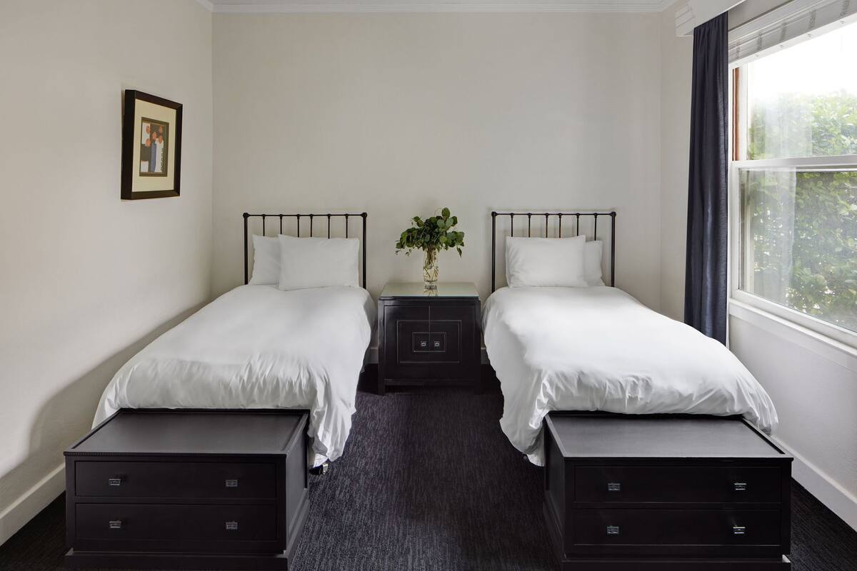 A twin room with two beds, a small bedside table with a vase, and a window with curtains.