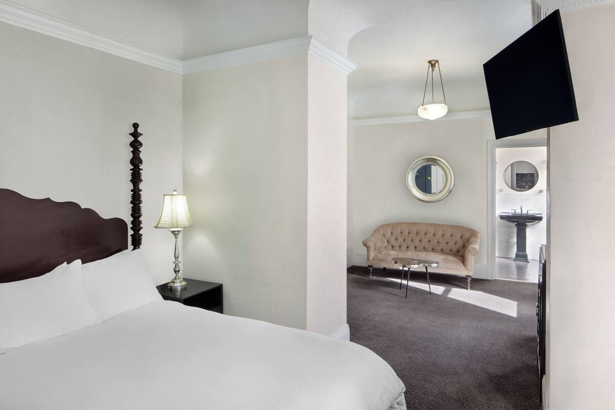 A hotel room with a large bed, a nightstand with a lamp, a sofa, a round mirror, and a television mounted on the wall.