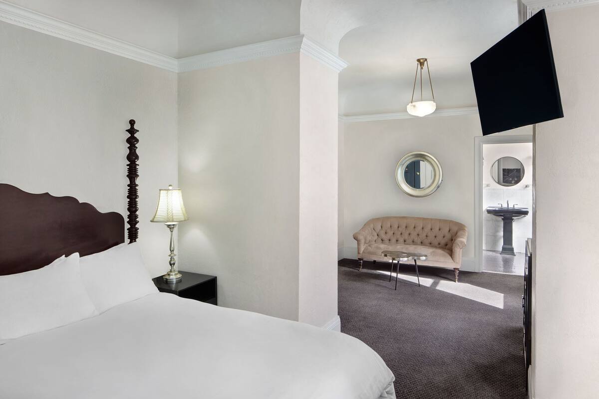 A hotel room with a large bed, a nightstand with a lamp, a beige sofa, a round mirror, and a television mounted on the wall.