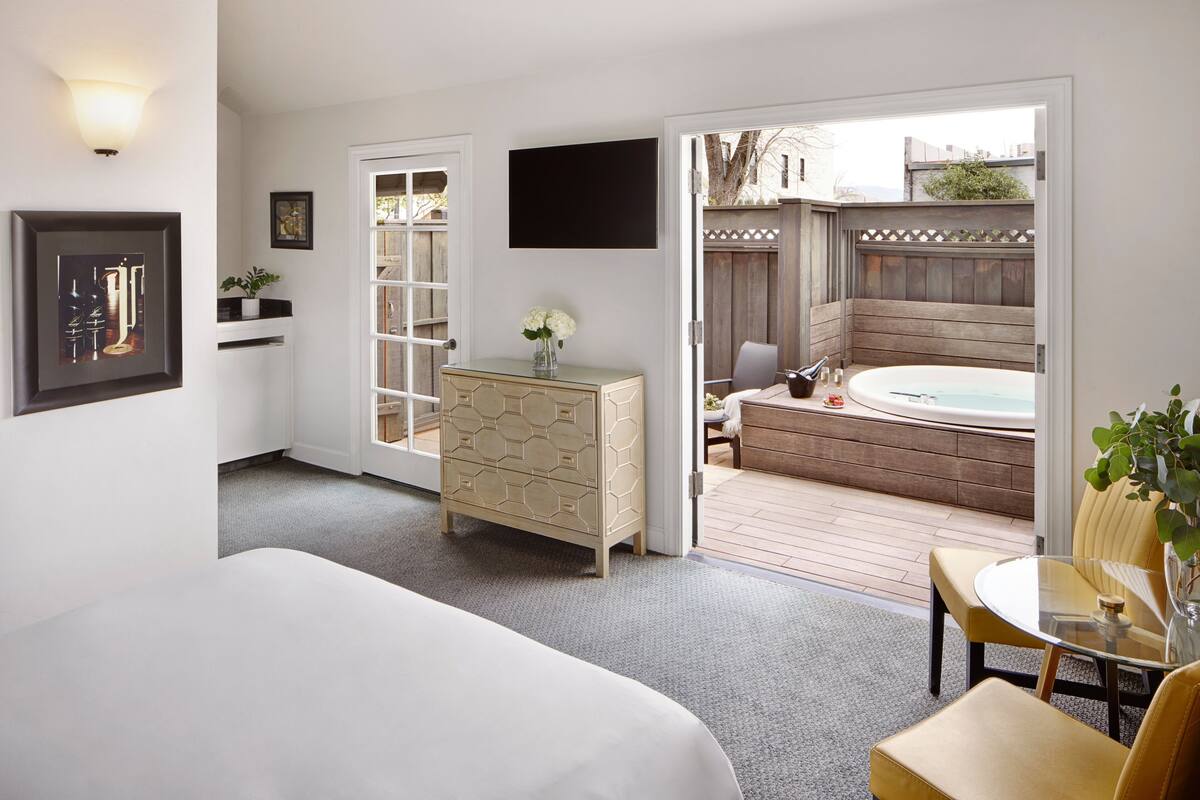 A hotel room with a bed, a dresser, a TV, and a view of an outdoor hot tub and deck.