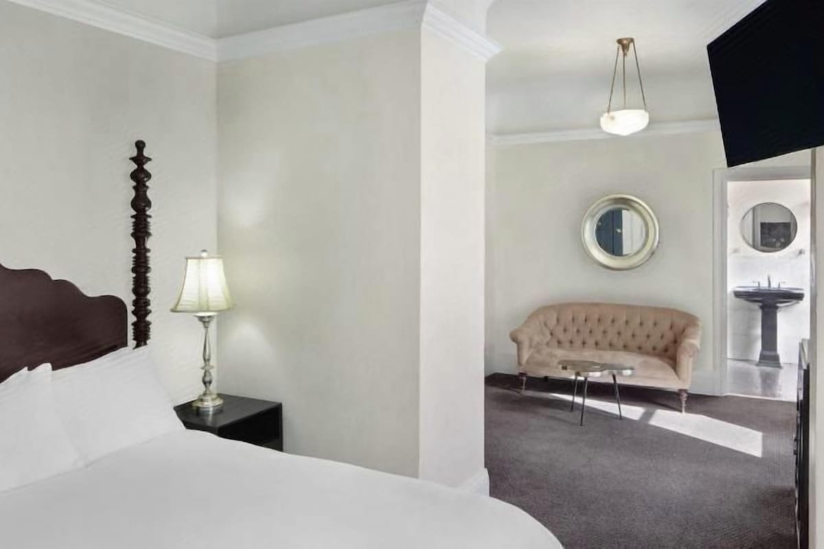 A hotel room with a bed, a nightstand with a lamp, a sofa, a round mirror, and a television mounted on the wall.