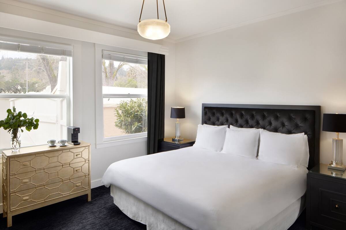 A hotel room with a large bed, a bedside table with a lamp, a dresser, and a window with a view of the outdoors.