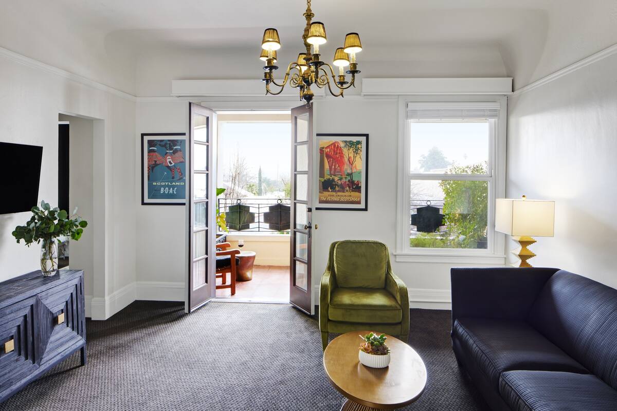 A modern living room with a sofa, a green armchair, a coffee table, and a chandelier. There are two windows with artwork and a door leading to another room.