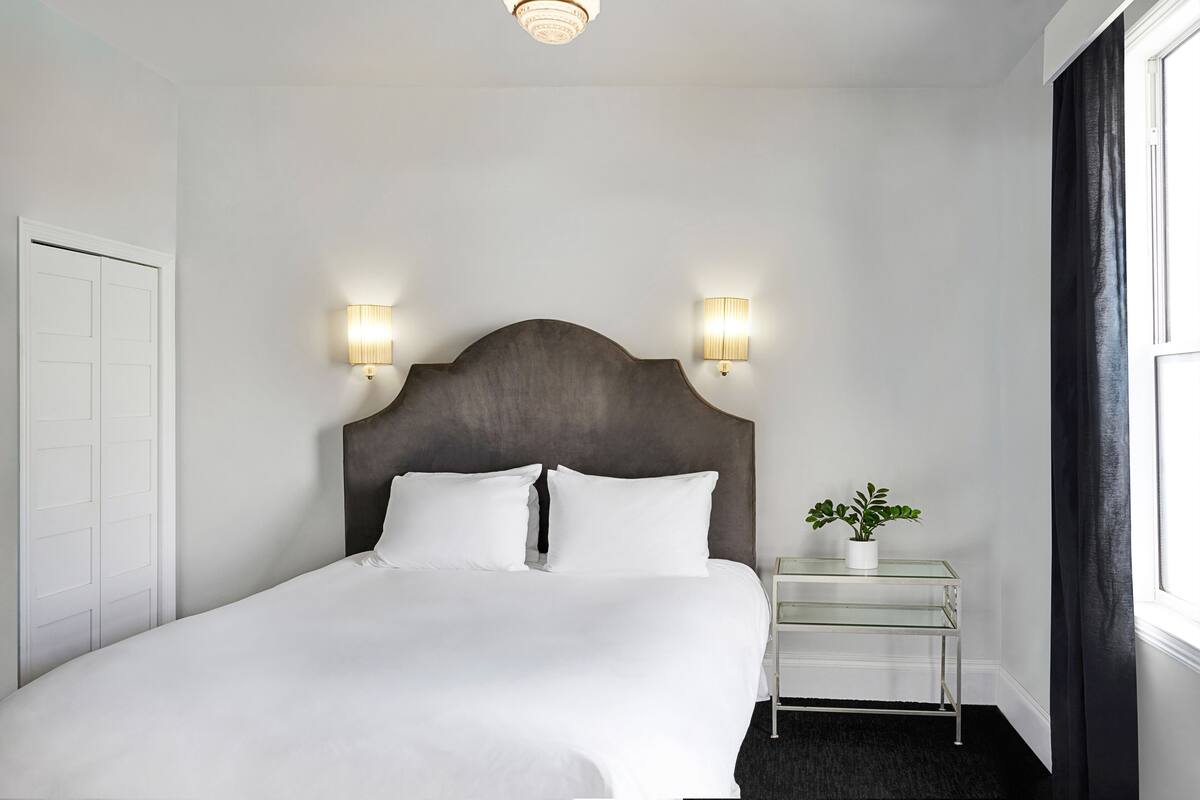 A neatly made bed with white linens, a dark headboard, two wall-mounted lamps, and a small bedside table with a plant.