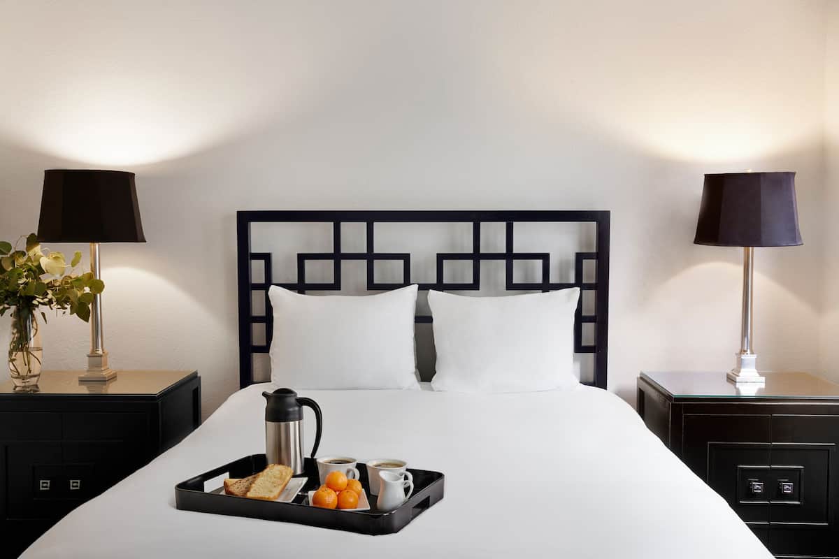A neatly made bed with a tray of breakfast items, including bread, oranges, and coffee, in a hotel room with a modern black headboard and two bedside lamps.