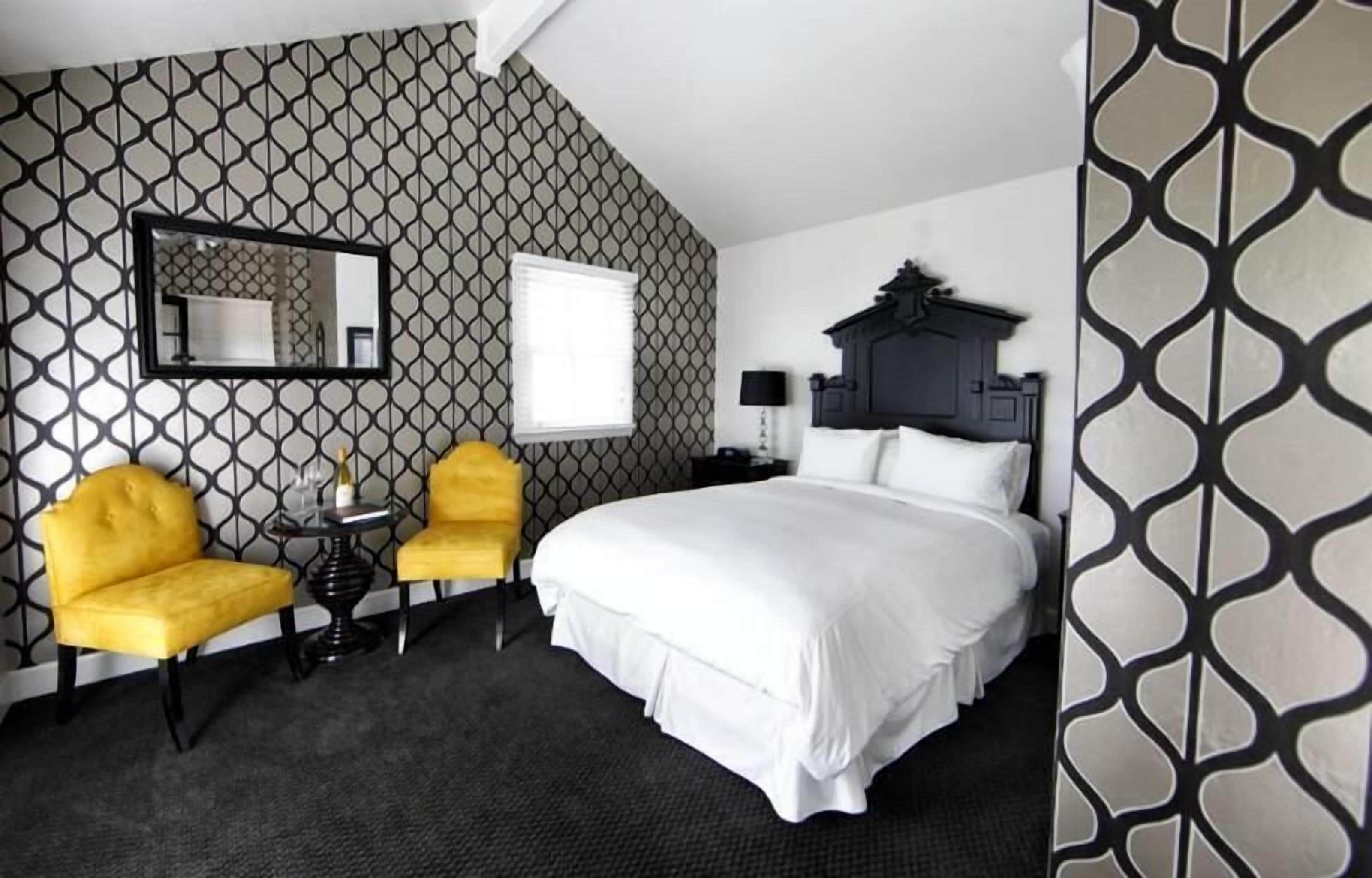 Une chambre d’hôtel avec un grand lit, deux fauteuils jaunes, une petite table, un miroir et un mur décoré de motifs.