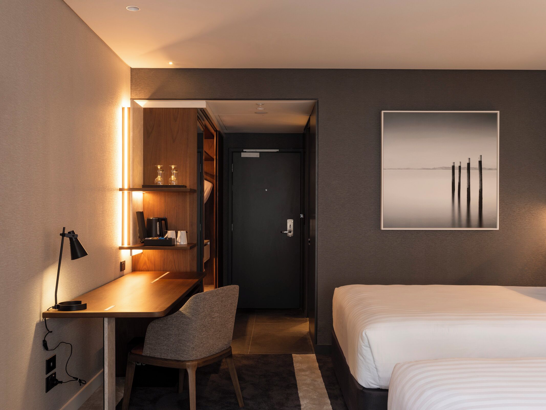 A hotel room with a bed, a desk with a chair, a bedside table with a lamp, a framed picture on the wall, and a door leading to a bathroom.