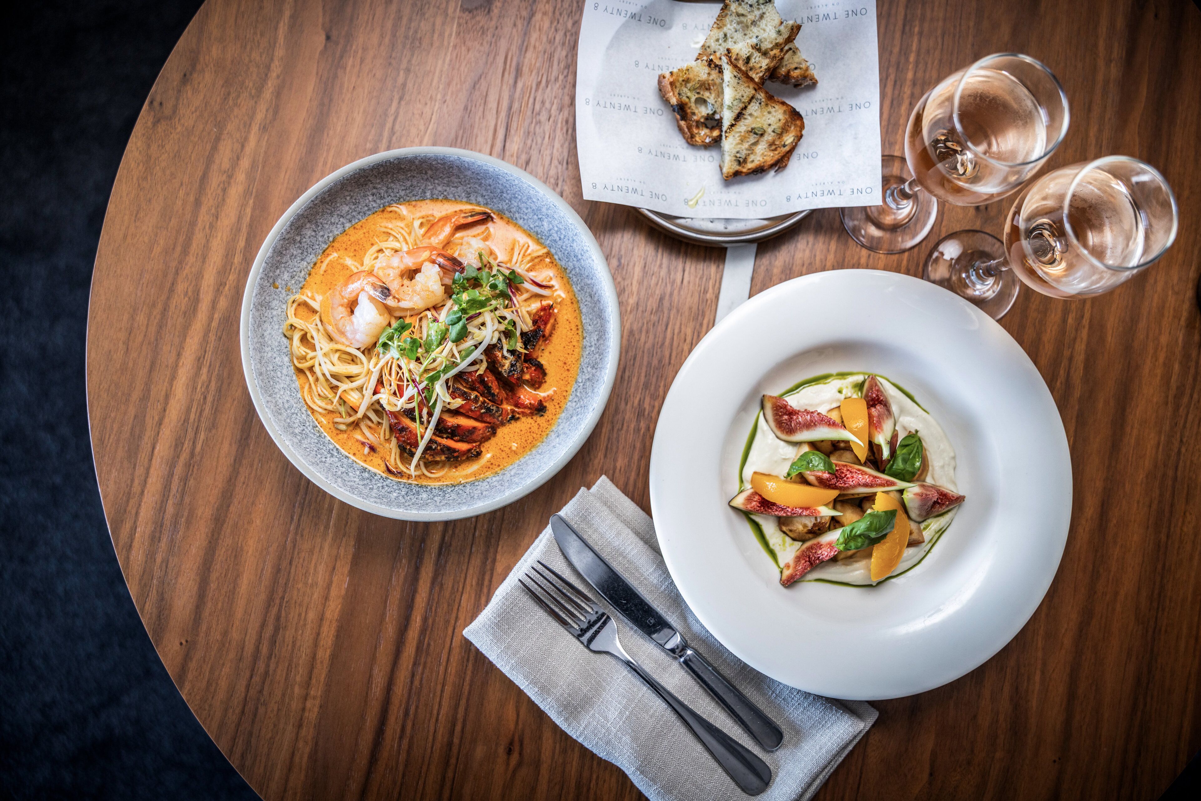 Una mesa con un plato de pasta, una ensalada, copas de vino y cubiertos.