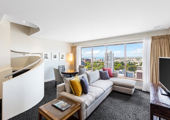 A modern hotel room with a curved staircase, a large window with a city view, a sofa with cushions, a coffee table, and a flat-screen TV.