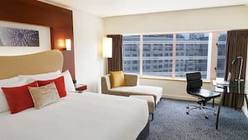 A hotel room with a large bed, a desk, a chair, and a window with a city view.