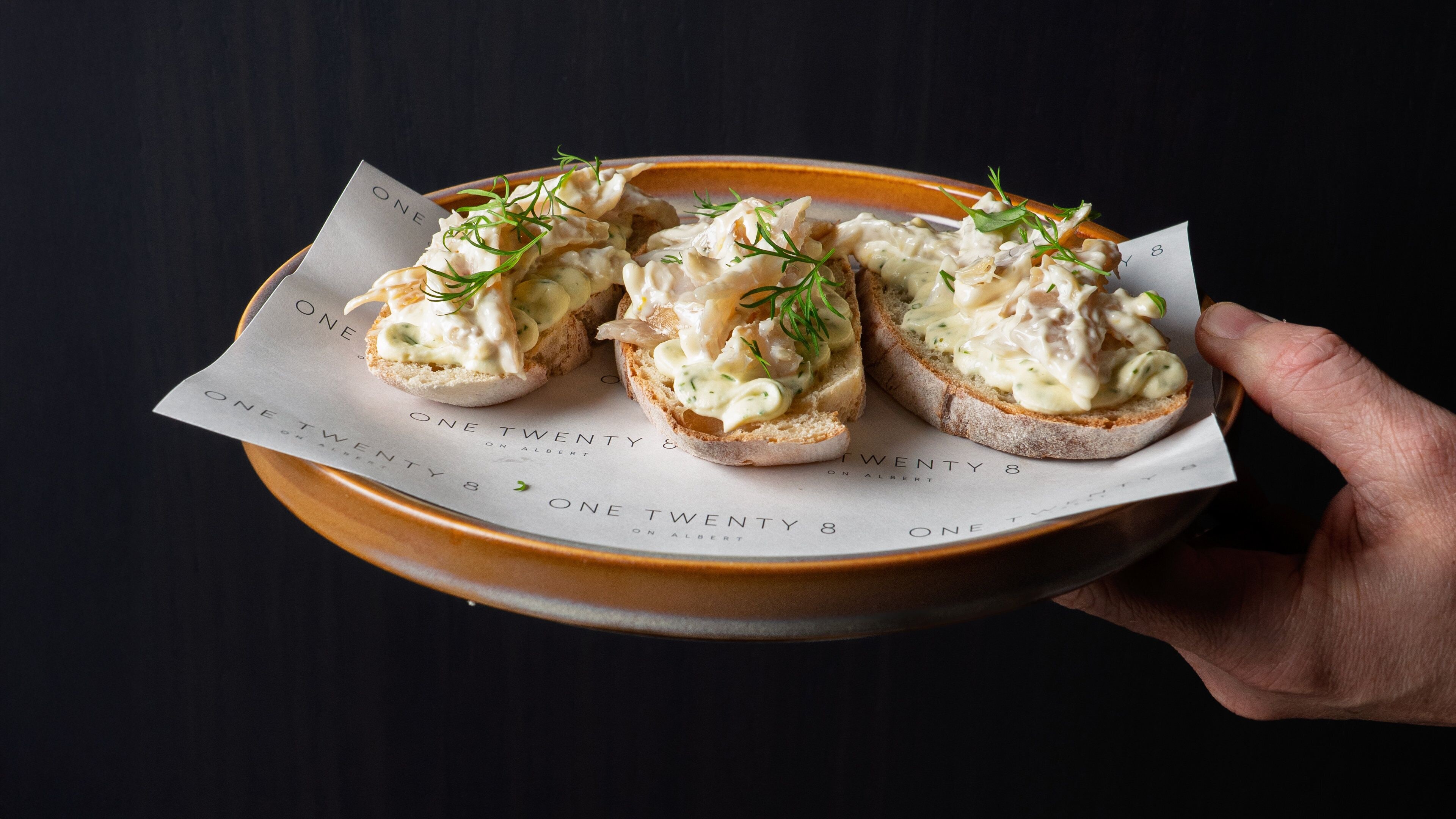 A hand holding a plate with three open-faced sandwiches on a paper with text.
