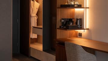 Habitación de hotel con escritorio de madera, una silla, bata colgada en un perchero y una estantería con cafetera y vasos.