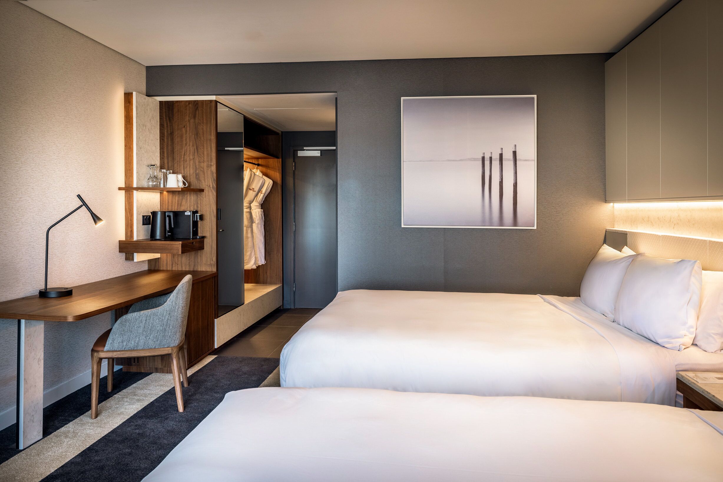 A hotel room with two beds, a desk, a chair, a wardrobe, and a framed picture on the wall.