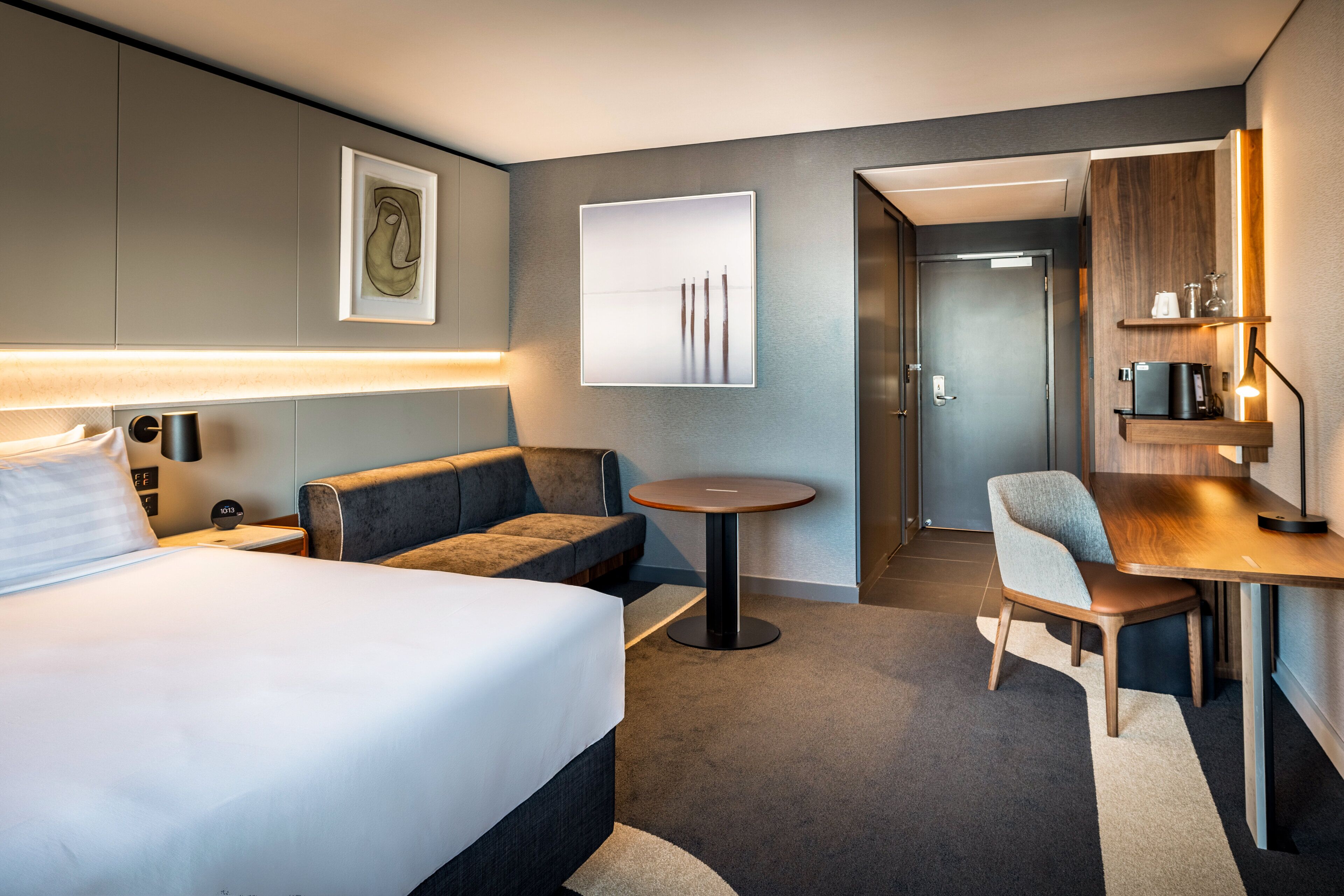 A hotel room with a bed, a sofa, a small round table, a desk, a chair, a lamp, and artwork on the walls.