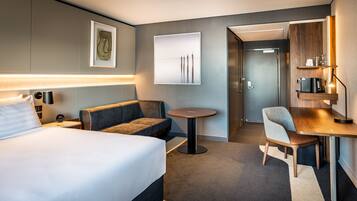 A hotel room with a bed, a sofa, a small round table, a desk, a chair, a lamp, and artwork on the walls.