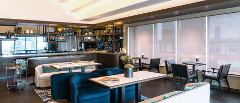 A modern hotel lobby with a bar, seating area, and large windows offering city views.