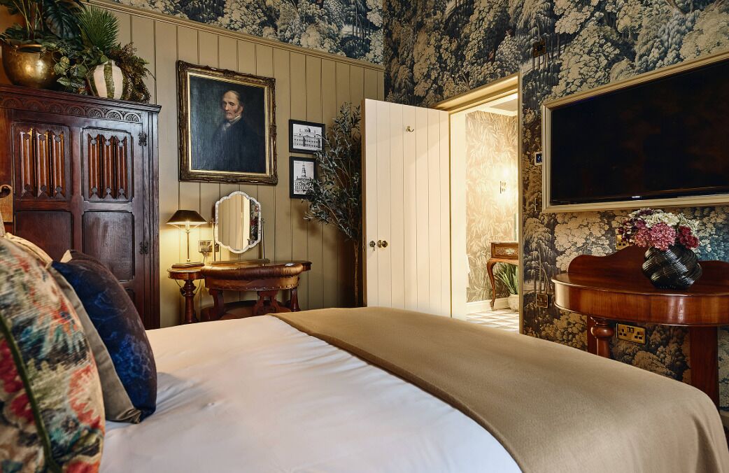 A bedroom with a bed, a dresser, a mirror, a table with a vase, and a door leading to another room.