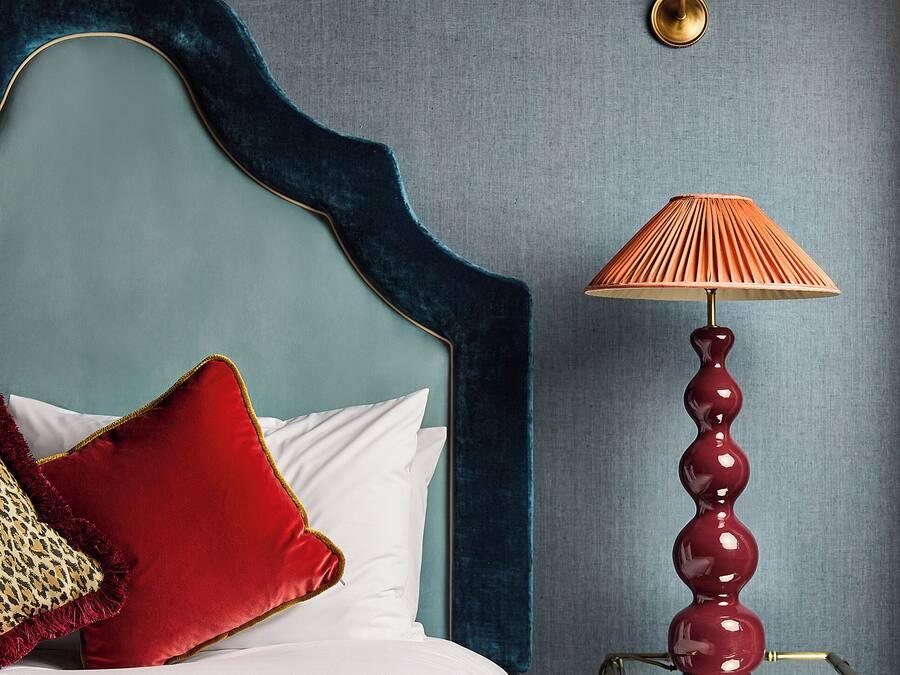A bed with a blue headboard, red and patterned pillows, a white comforter, and a red lamp on a bedside table.