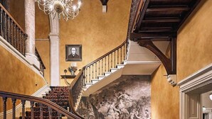 A grand staircase with ornate railings and patterned carpet, leading to a room with a mural and a chandelier. - Brownsover Hall Hotel  (Rugby)