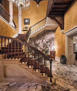 A grand staircase with ornate railings and patterned carpet, leading to a room with a mural and a chandelier. - Brownsover Hall Hotel  (Rugby)