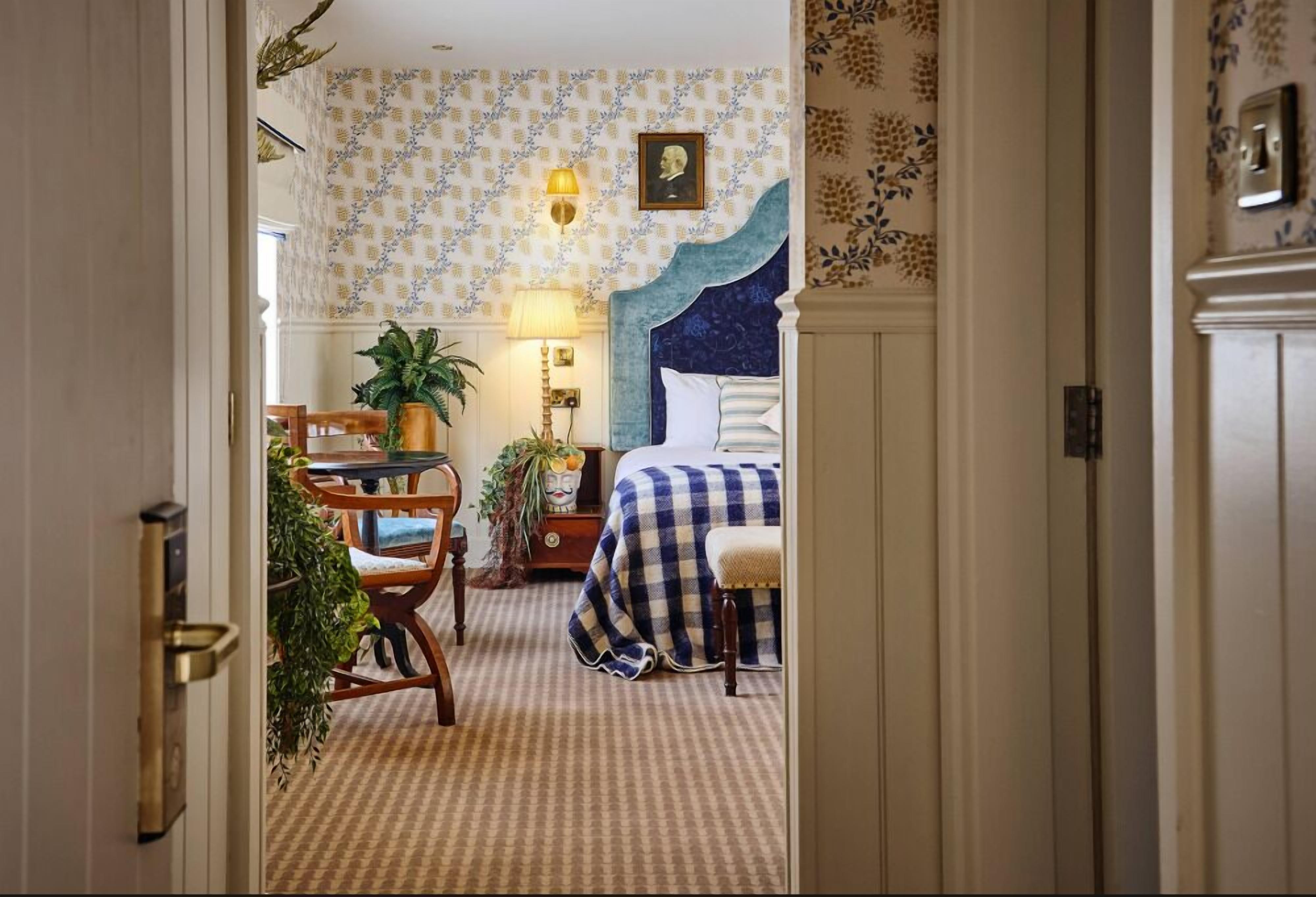 A bedroom with a checkered bedspread, a bedside table with a lamp, a chair, a potted plant, and a wall-mounted picture.