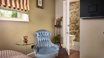 A room with a tufted armchair, a small glass table with a vase, a bed, a window with curtains, a door leading to a bathroom, and a television on the wall.