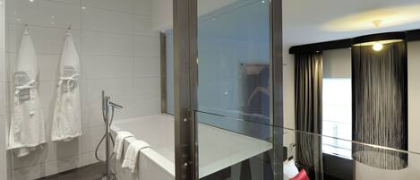 Una habitación de hotel moderna con separador de vidrio, una cama grande con ropa de cama roja y blanca, un box de ducha de vidrio y vistas a la ciudad por la ventana.