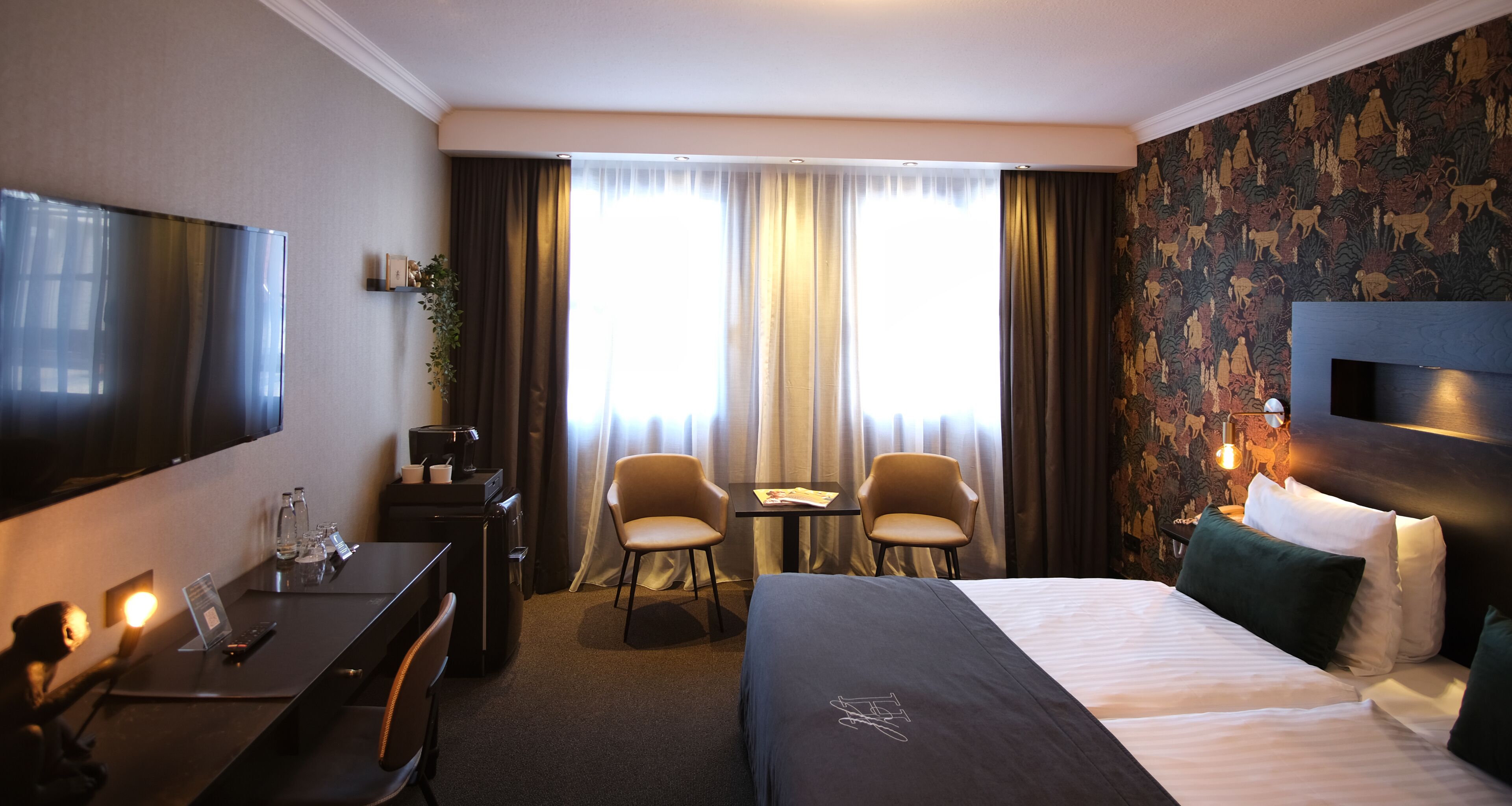 A hotel room with a bed, desk, chairs, a TV, and a window with curtains.