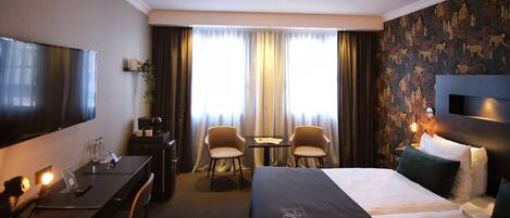 Una habitación de hotel con cama, escritorio, sillas, televisor y ventana con cortinas.
