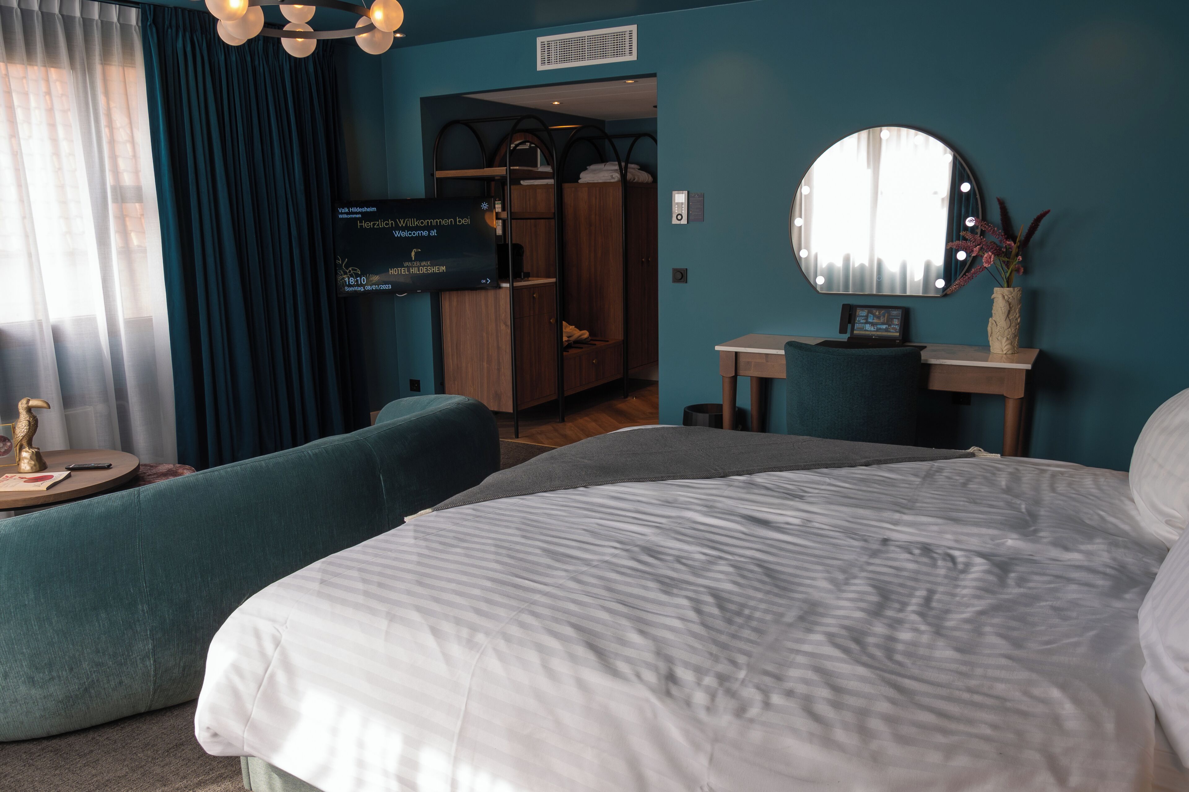 A hotel room with a large bed, a seating area, a vanity with a mirror, and a wardrobe.