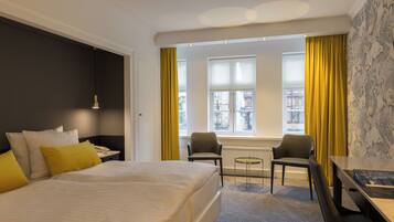 A hotel room with a bed, two chairs, a desk, and a view of buildings through the window.