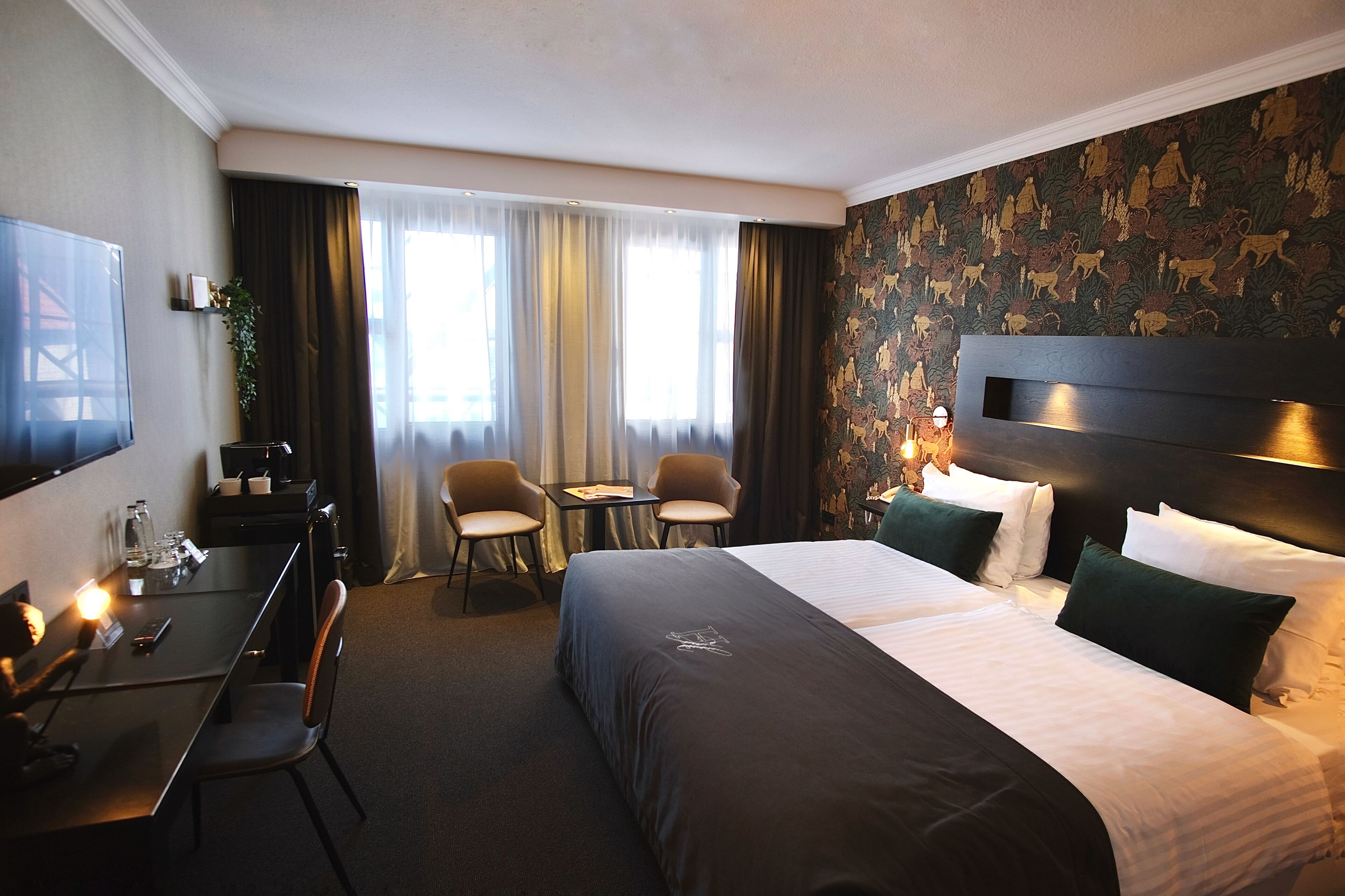A hotel room with a bed, two chairs, a desk, a TV, and a window with curtains.