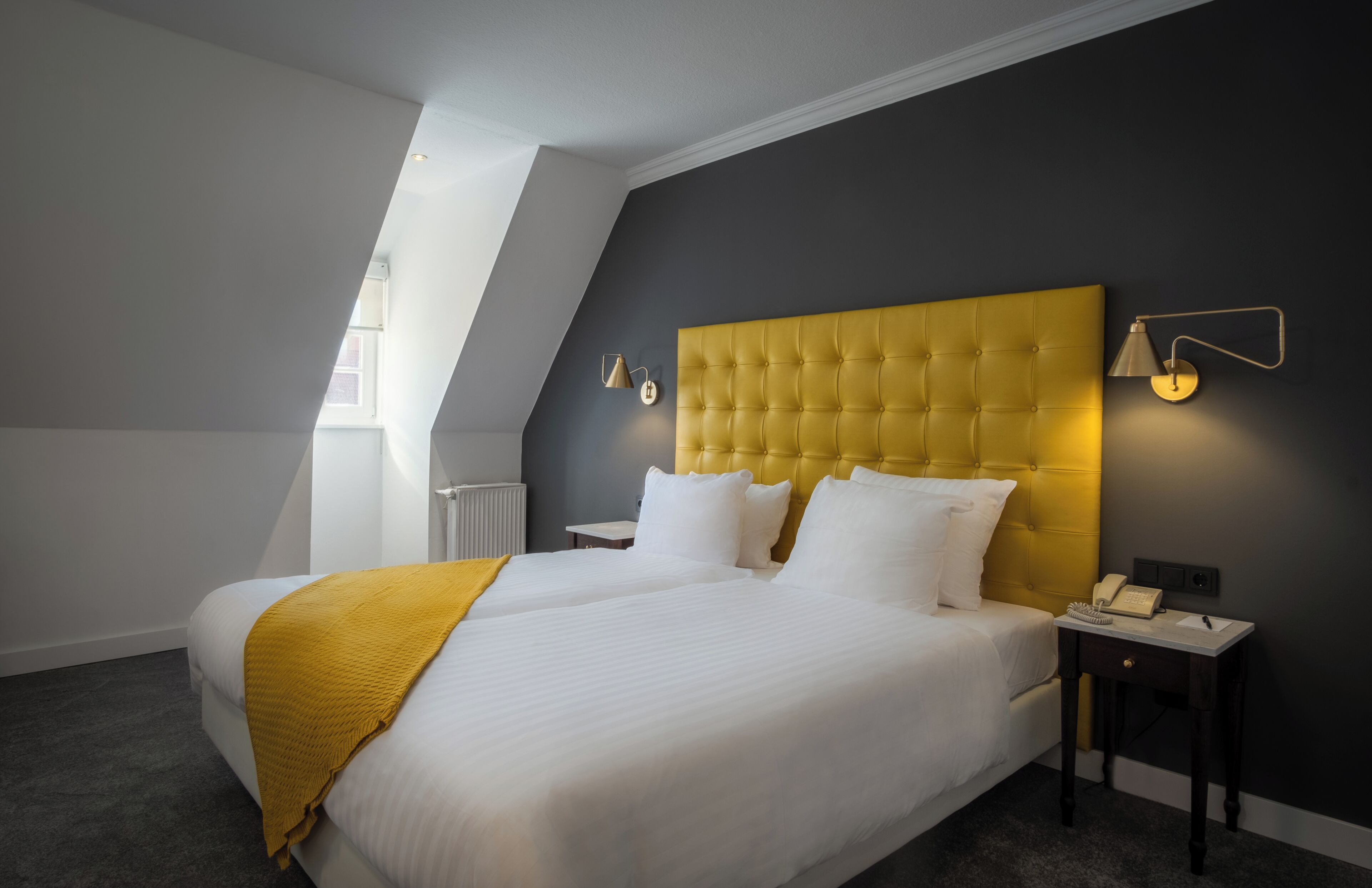Ein Hotelzimmer mit einem großen Bett, einem gelben Kopfteil, einem Nachttisch mit Lampe und Blick auf den Himmel durch ein Oberlicht.