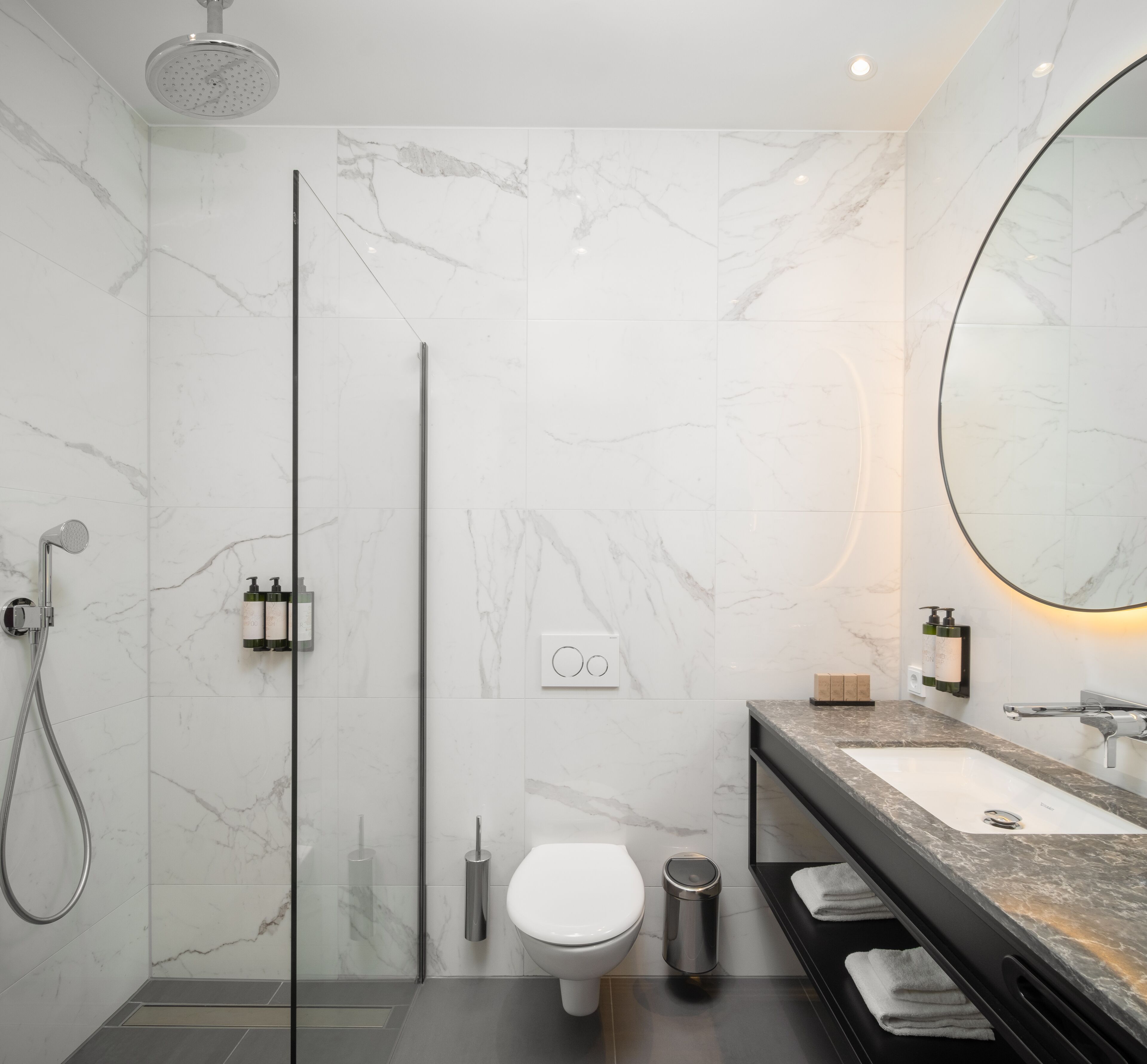 A modern bathroom with a glass shower enclosure, a toilet, a sink with a marble countertop, and a large mirror.