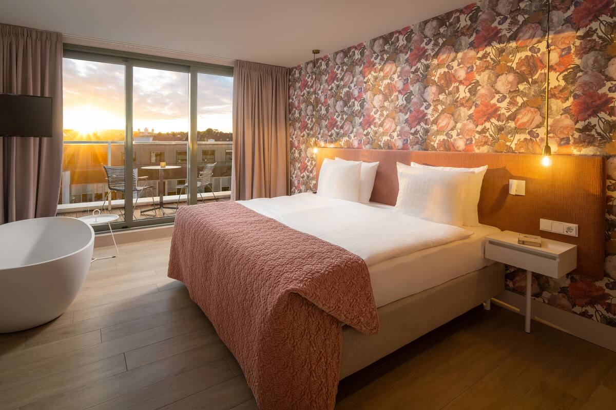 Une chambre d’hôtel avec un grand lit, une table de chevet blanche, une commode, une grande baignoire blanche, un balcon avec vue sur le coucher de soleil et un papier peint à motifs floraux.