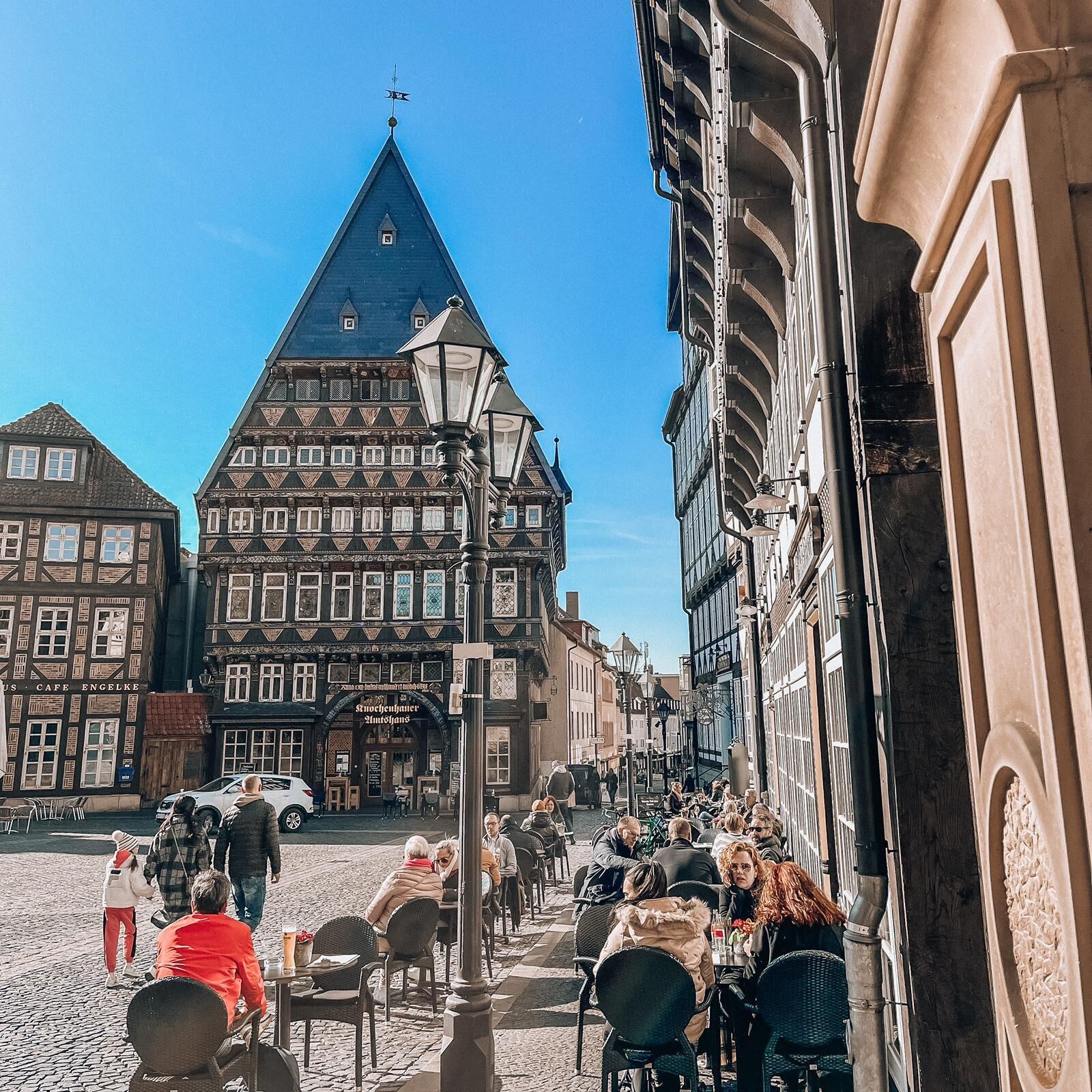 A cobblestone street with traditional European architecture, including a prominent building with a steeply pitched roof and a smaller building with a sign reading "Cafe Engelke".