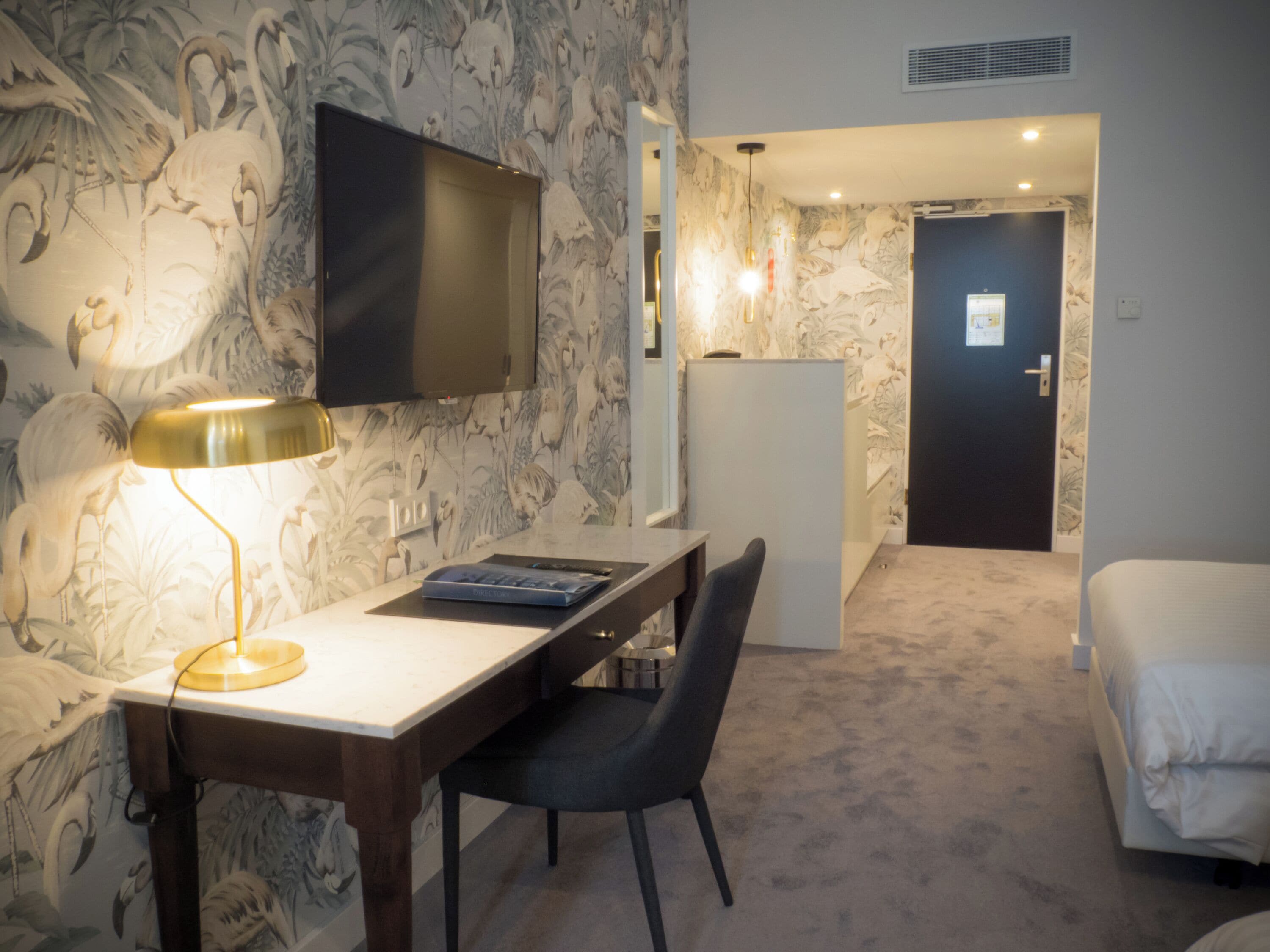 Ett hotellrum med ett skrivbord, en stol, en säng, en tv och en dörr som leder till ett annat rum.