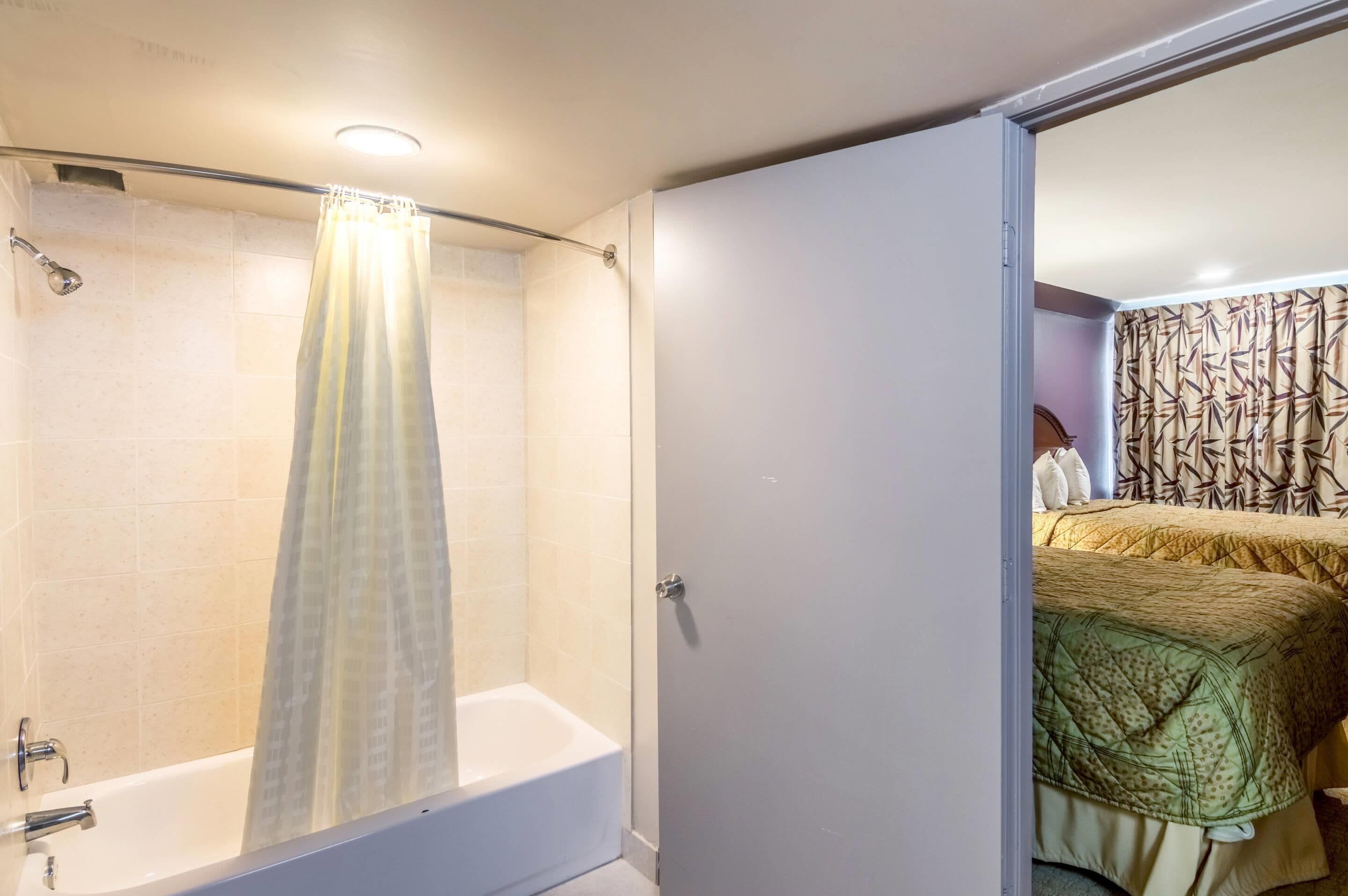 Une salle de bain avec une baignoire, un rideau de douche et une porte visible donnant sur une chambre à coucher où se trouve un lit.